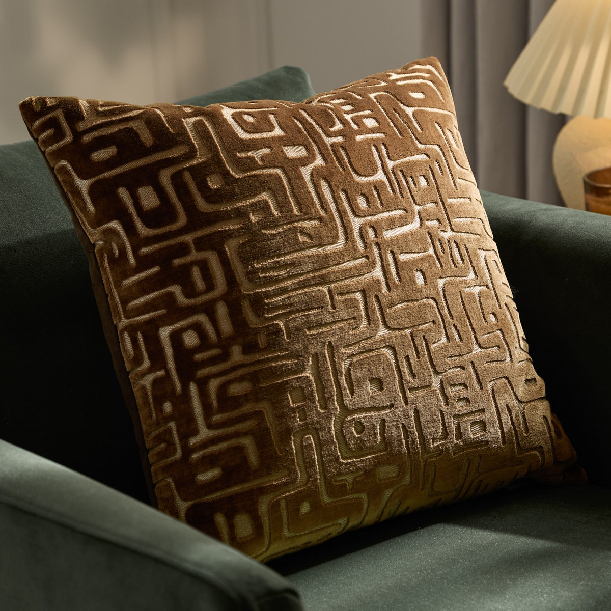 Soren Abstract Velvet Cushion in brown, square with a raised cut-velvet geometric maze pattern shifting from dark brown to soft gold, shown on a green sofa.