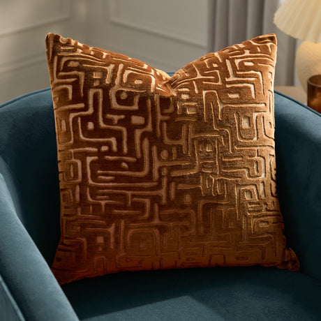 Soren Abstract Velvet Cushion in copper, showing a maze-like cut velvet pattern in burnt orange tones, styled on a teal armchair.