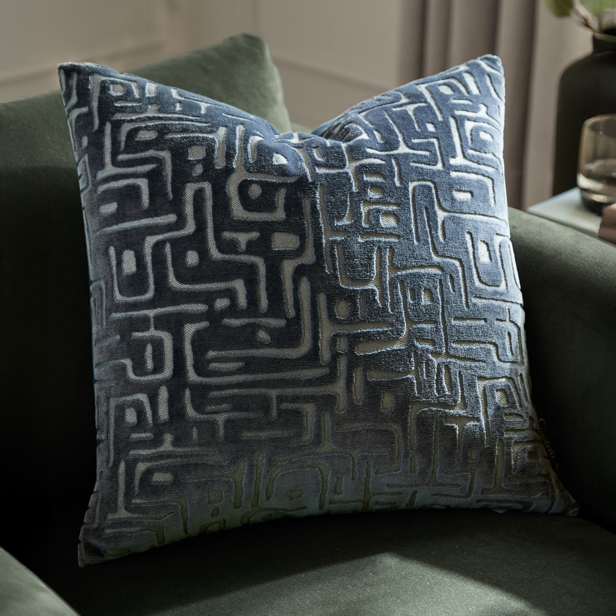 Soren Abstract Velvet Cushion in slate blue, a 45cm square cushion with a geometric maze-like cut velvet pattern, shown on a green upholstered armchair.