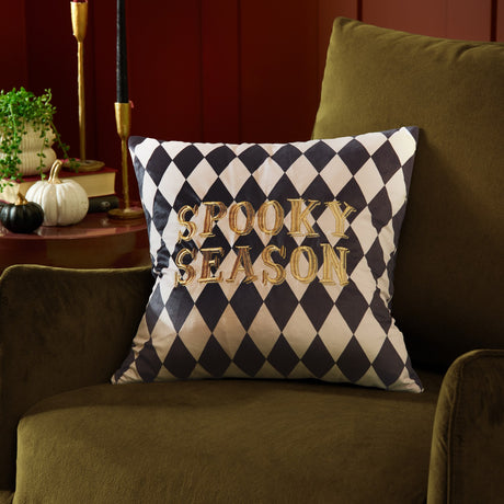 Spooky Season Geometric Velvet Cushion in black and white harlequin print with raised gold "Spooky Season" embroidery, shown on an olive velvet armchair in a living room setting.