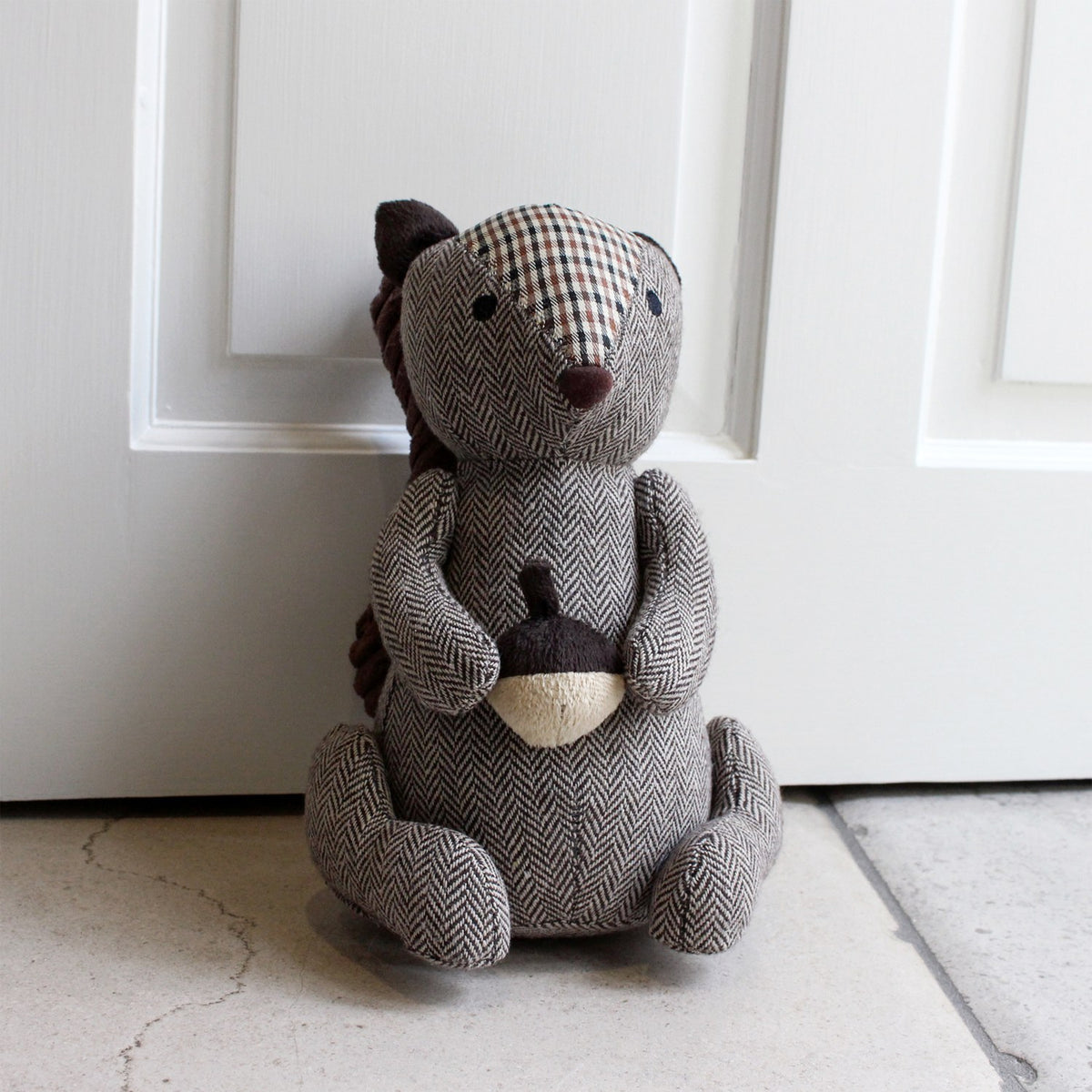 Squirrel Acorn Polyester Door Stop in brown, sitting upright holding an acorn, with herringbone body, tartan-patch head, and ribbed tail, shown on a hallway floor by a white door.