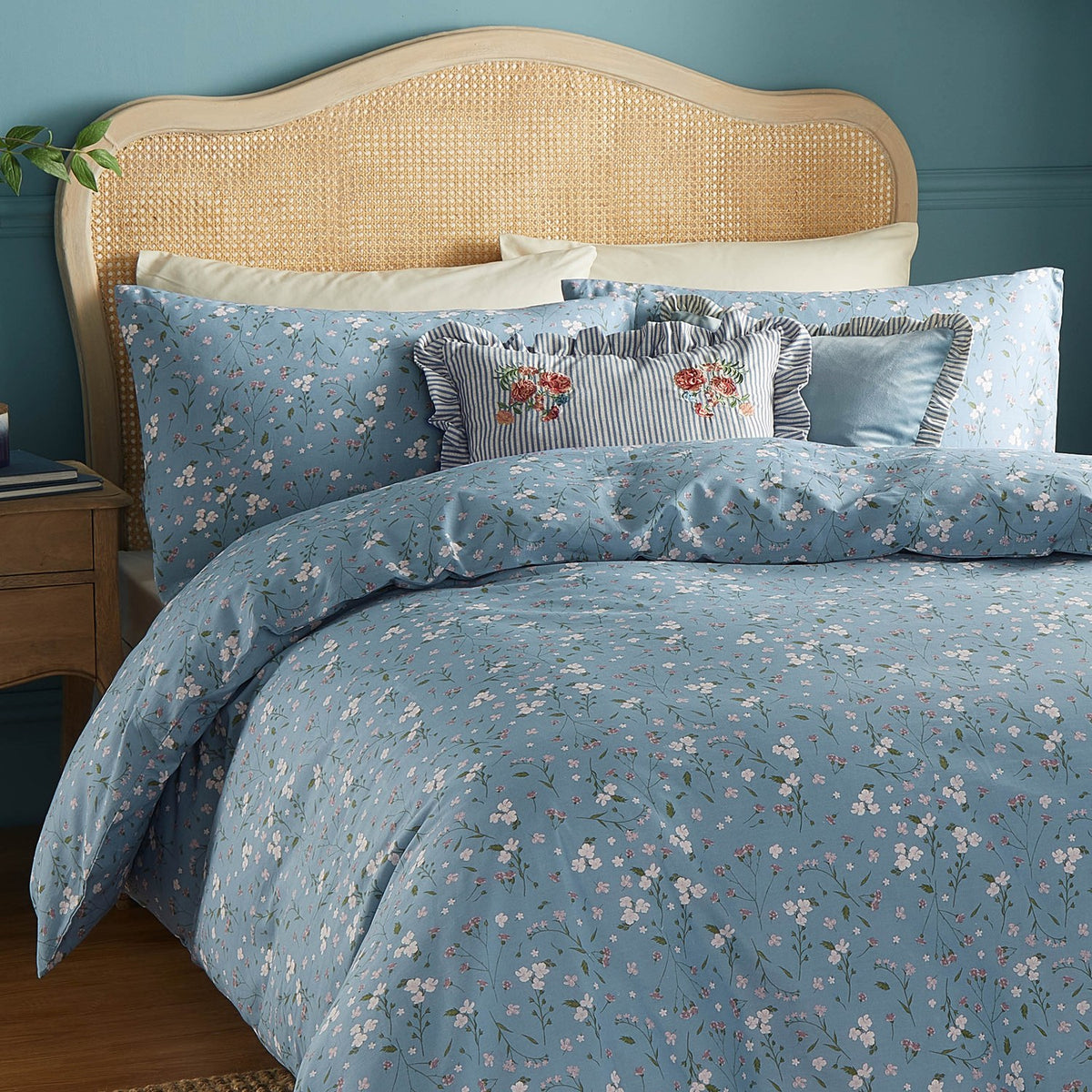 Solene Ruffle Velvet Linen Cushion in dusky blue with striped linen-blend ruffle edge, shown on a floral blue bedspread against a rattan headboard.