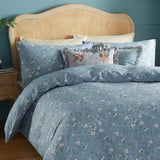 Solene Ruffle Velvet Linen Cushion in dusky blue with striped linen-blend ruffle edge, shown on a floral blue bedspread against a rattan headboard.