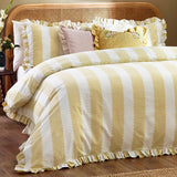 Solene Ruffle Velvet Linen Cushion in pink on a striped yellow and white bed, square velvet cushion with gathered ruffle trim, styled among patterned pillows.