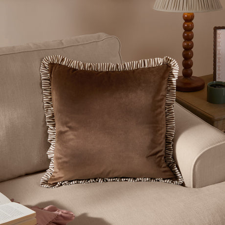 Solene Ruffle Velvet Linen Cushion in truffle brown with striped ruffle trim, matt velvet face, shown on a beige sofa beside a lamp table.