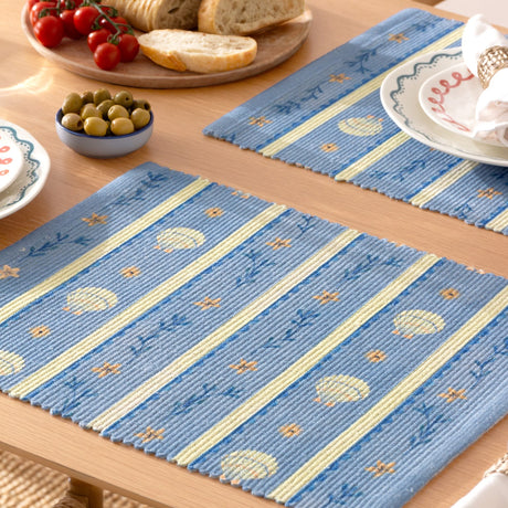 Blue polyester placemats with printed scallop shell, starflower and sprig design in striped bands, shown on a wooden dining table with tableware.