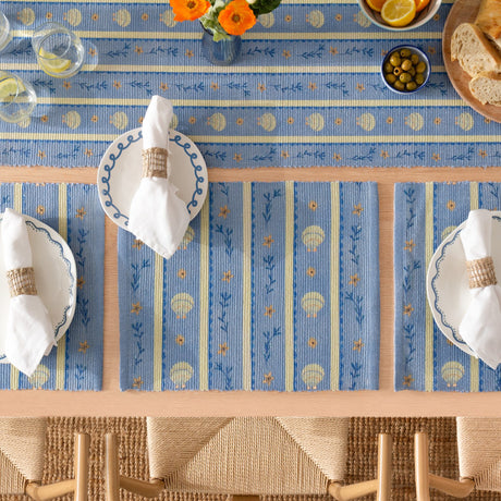 Sea Shells printed polyester placemats in blue with cream stripes and shell motifs, set on a dining table with white plates, linen napkins and rope napkin rings.