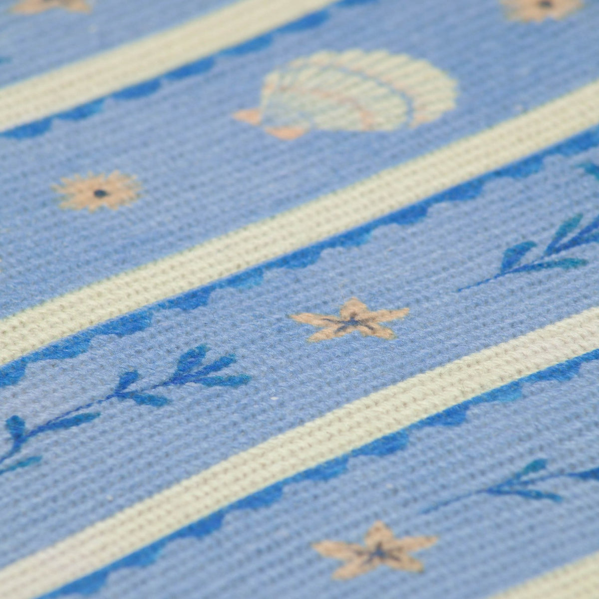 Close-up of a Sea Shells printed polyester placemat in blue, showing scallop shells, starfish and sprig motifs between cream woven stripes.