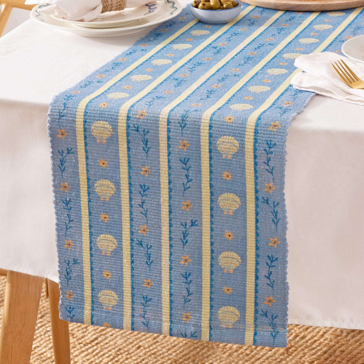 Blue polyester table runner with printed scallop shells, flowers and leafy sprigs between pale yellow stripes, styled on a dining table with plates and cutlery.