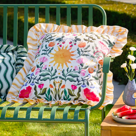Summer Solstice square cushion with sun, floral and striped ruffled edge print in cream, yellow and multicoloured tones, shown on a green outdoor bench.