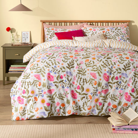 Summer Solstice reversible duvet cover set on a bedroom bed, floral print in pink, orange, lilac and coral on olive stems with matching pillowcases and amber wiggle-stripe trim.