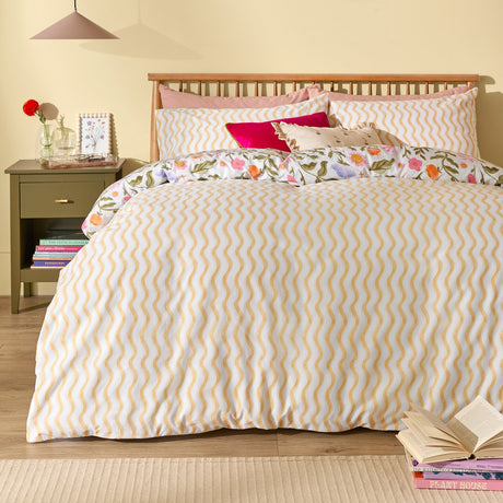 Summer Solstice reversible duvet cover set on a bedroom bed, amber and cream wiggle stripe side shown, with floral pillowcases in pink, orange and lilac visible beneath.