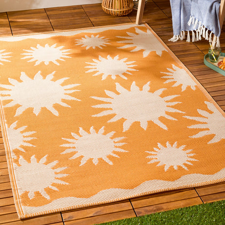 Summer Solstice reversible outdoor rug in sunrise yellow, featuring cream sun motifs on an orange ground, laid on a wooden deck beside outdoor furniture.