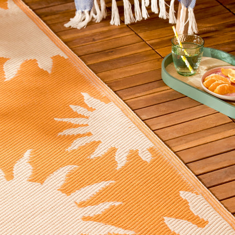 Summer Solstice Reversible Abstract Rug in Sunrise Yellow, orange and cream sunburst pattern, shown on a wooden deck beside a tray with a drink and fruit.