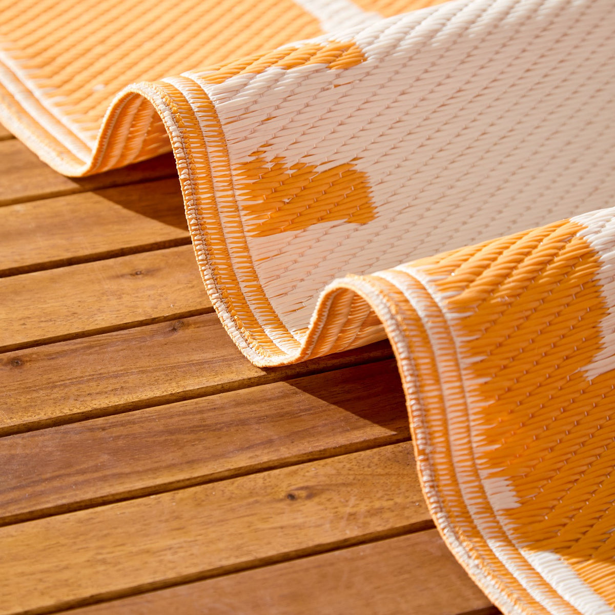 Summer Solstice Reversible Abstract Rug in Sunrise Yellow, folded to show woven polypropylene texture in orange and cream abstract pattern, shown outdoors on a wooden deck.