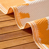 Summer Solstice Reversible Abstract Rug in Sunrise Yellow, folded to show woven polypropylene texture in orange and cream abstract pattern, shown outdoors on a wooden deck.
