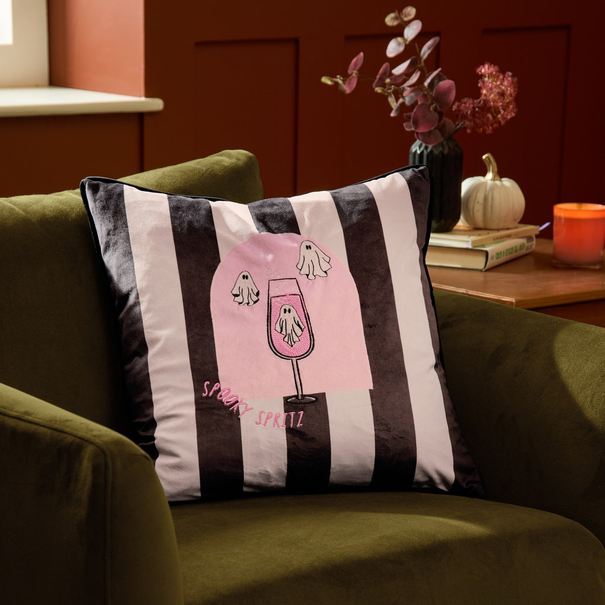 Spooky Spritz striped velvet cushion in pink and mono, featuring a ghost-topped cocktail glass design, shown on an olive green armchair in a living room setting.