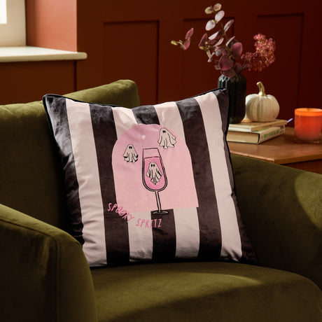 Spooky Spritz striped velvet cushion in pink and mono, featuring a ghost-topped cocktail glass design, shown on an olive green armchair in a living room setting.