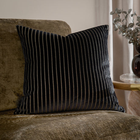 Savoy Stripe velvet cushion in black with fine gold-lined vertical stripes, shown on a khaki velvet armchair beside a side table and vase.