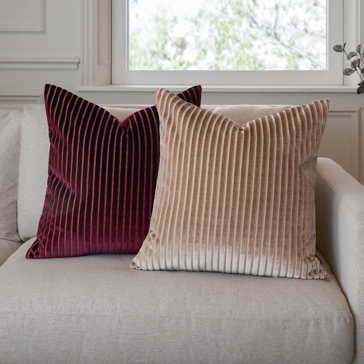 Savoy Stripe Polyester Velvet Cushion in burgundy with ribbed vertical pile, shown beside a matching beige version on a light sofa near a window.