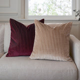 Savoy Stripe Polyester Velvet Cushion in burgundy with ribbed vertical pile, shown beside a matching beige version on a light sofa near a window.