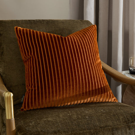 Savoy Stripe Polyester Velvet Cushion in brick orange, square with ridged, striped cut-velvet front, styled on a brown armchair beside curtains.