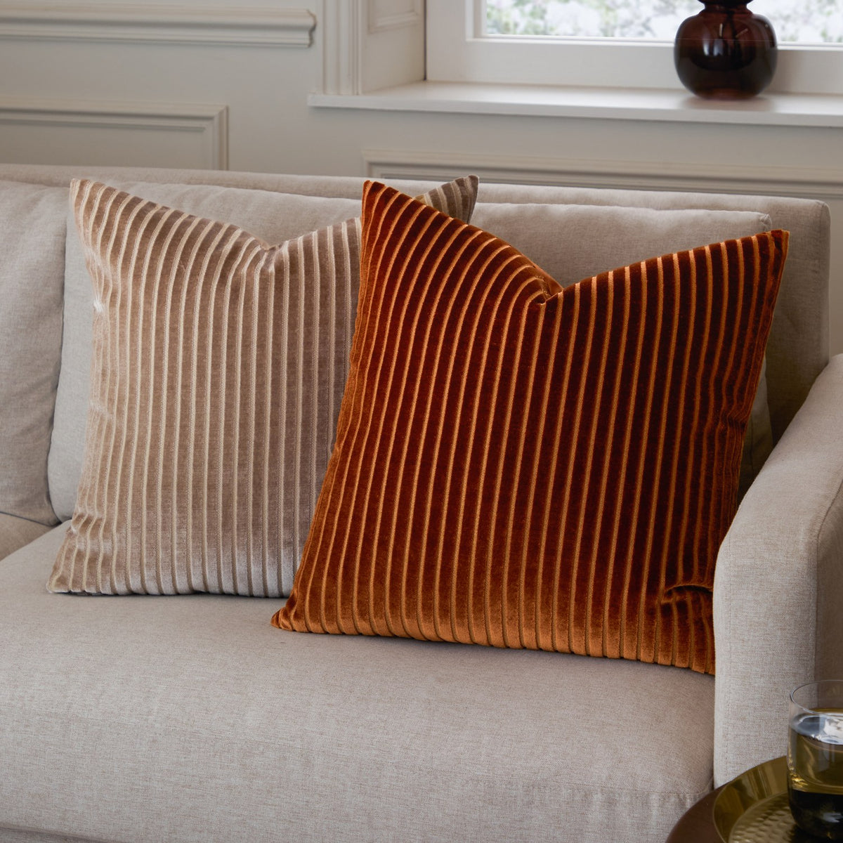 Savoy Stripe Polyester Velvet Cushion in Brick, a ribbed square cushion with vertical cut-velvet stripes, shown on a beige sofa beside a beige striped cushion.