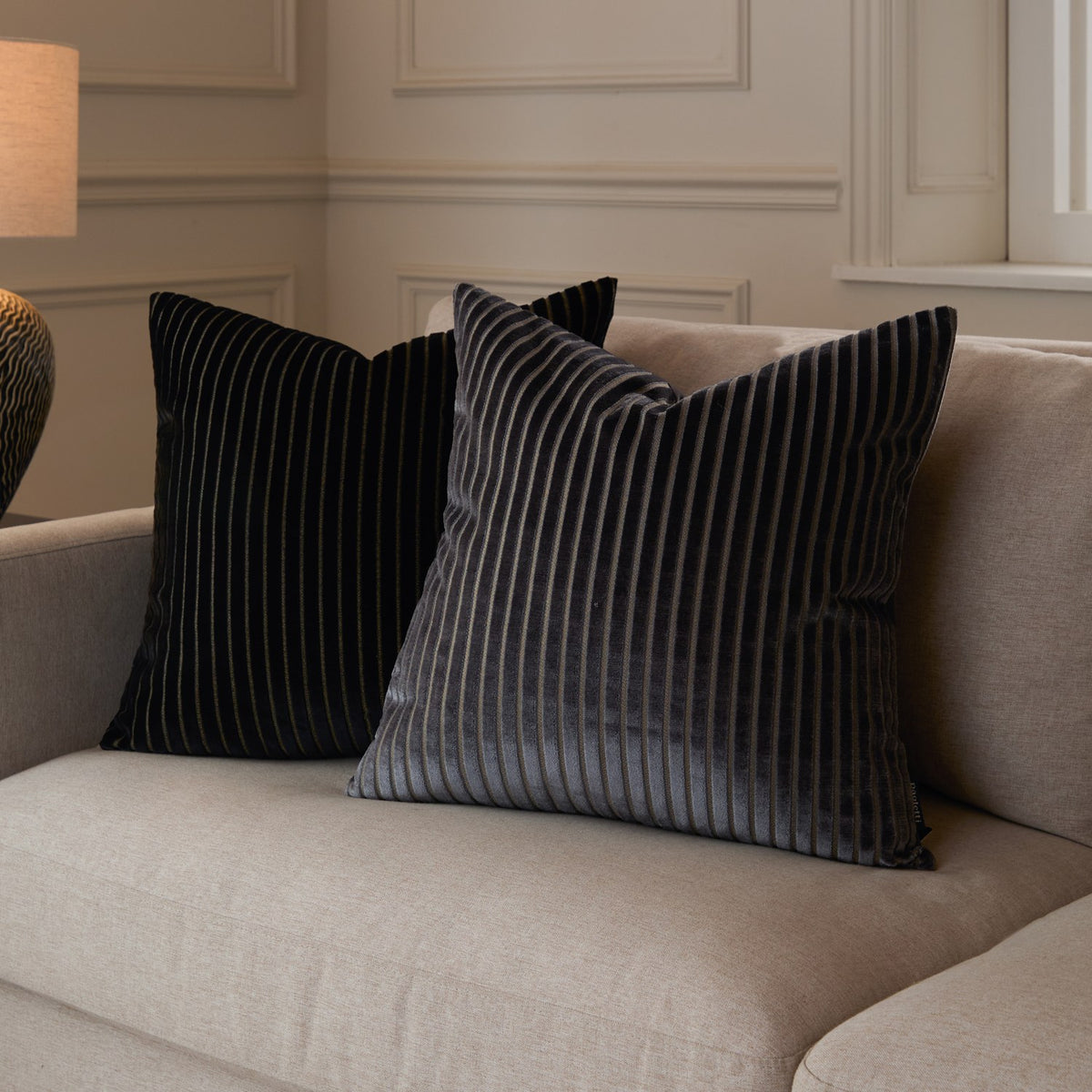 Savoy Stripe velvet cushion in charcoal grey with cut-pile stripe texture and fine metallic threads, styled on a beige sofa beside a matching black striped cushion.