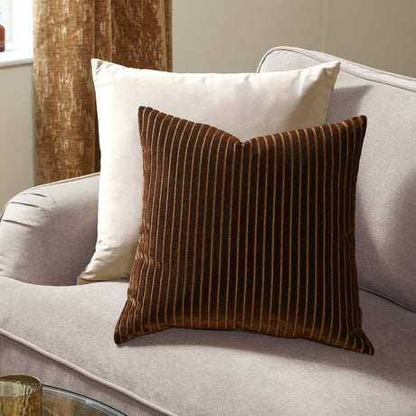 Savoy Stripe Polyester Velvet Cushion in chocolate brown with fine vertical amber-toned ridges, shown on a beige sofa alongside a cream cushion.