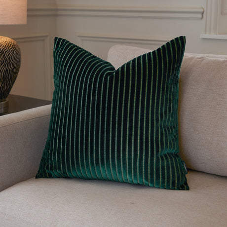 Savoy Stripe Polyester Velvet Cushion in emerald green with raised cut-velvet stripes, shown on a beige sofa in a living room setting.
