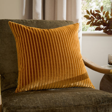 Savoy Stripe Polyester Velvet Cushion in gold with ribbed cut-velvet stripes, shown on a dark green armchair beside a window in a living room setting.