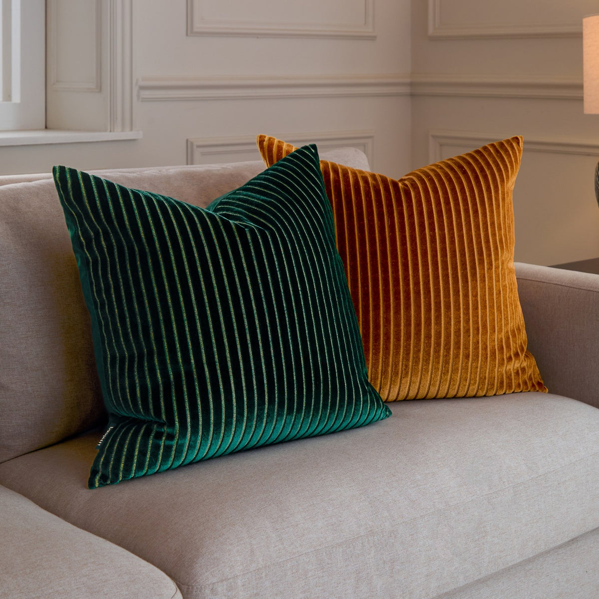 Savoy Stripe square cushion in deep gold ribbed velvet, shown beside a green striped cushion on a light beige sofa in a living room setting.