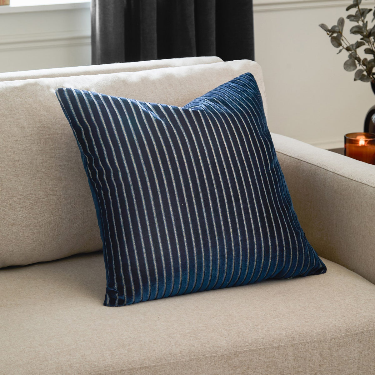 Savoy Stripe Polyester Velvet Cushion in navy with fine vertical stripes on a cut velvet front, shown on a cream sofa in a living room setting.