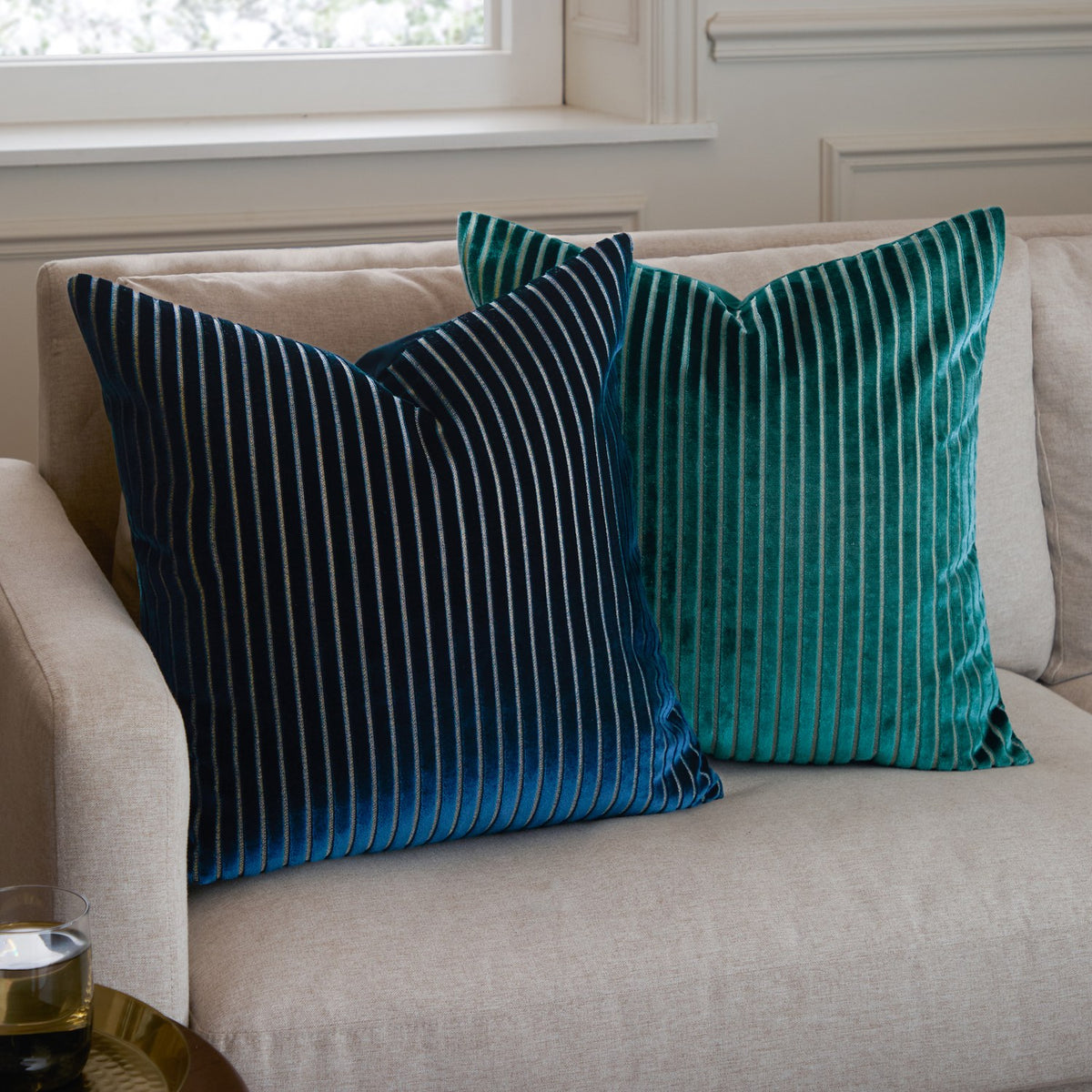 Savoy Stripe velvet cushion in navy with fine vertical striped front, shown 50x50cm square on a beige sofa beside a matching teal striped cushion.