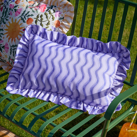 Rectangular outdoor lumbar cushion with cobalt blue wiggly stripe pattern on a pale lilac-white ground, ruffled edge trim, shown on a green garden bench.