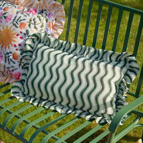 Summer Solstice Wiggly Stripe rectangular outdoor cushion in green wavy stripes with off-white ground and ruffled edge, shown on a green garden bench.