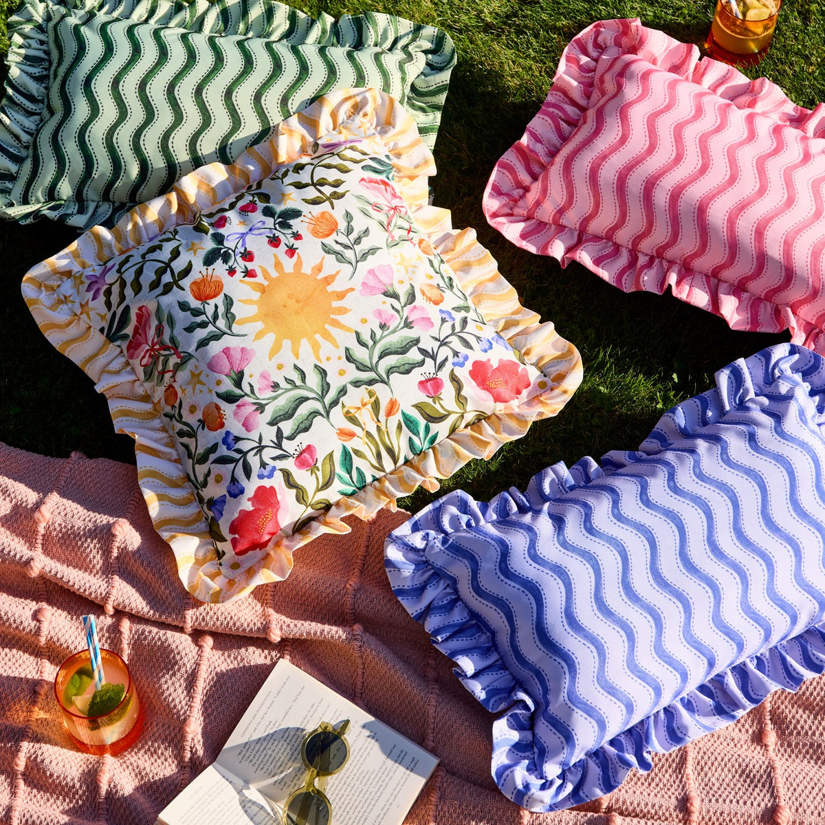 Green wiggly stripe rectangular cushion with ruffled edge, shown outdoors on grass alongside pink and blue striped cushions and a floral print cushion.