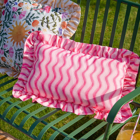 Summer Solstice Wiggly Stripe rectangular outdoor cushion in hot pink, wavy vertical stripe print with gathered ruffle trim, shown on a green garden bench.