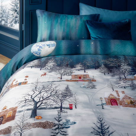 Starlight Cotton Piped Duvet Cover Set in Midnight Blue, printed with a snow-covered village scene, full moon and teal sky, styled on a bed with matching teal and navy pillows.