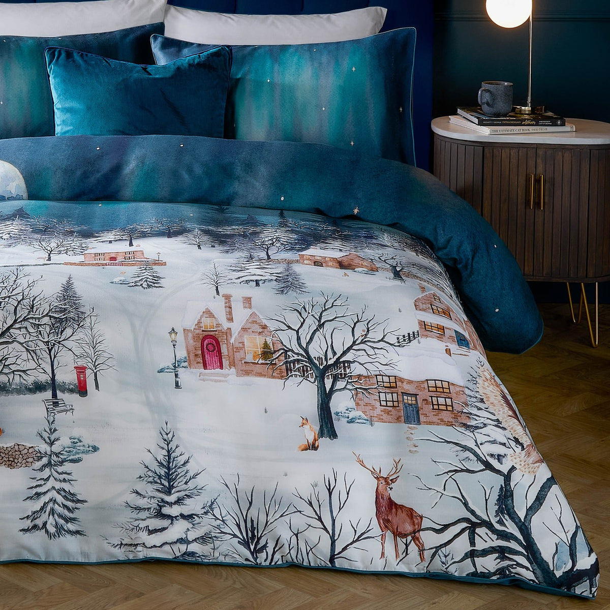 Starlight Cotton Piped Duvet Cover Set in midnight blue, shown on a bedroom bed with teal velvet cushions, printed with a snow-covered village scene featuring cottages, bare trees, a stag and fox.