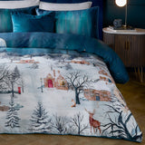 Starlight Cotton Piped Duvet Cover Set in midnight blue, shown on a bedroom bed with teal velvet cushions, printed with a snow-covered village scene featuring cottages, bare trees, a stag and fox.