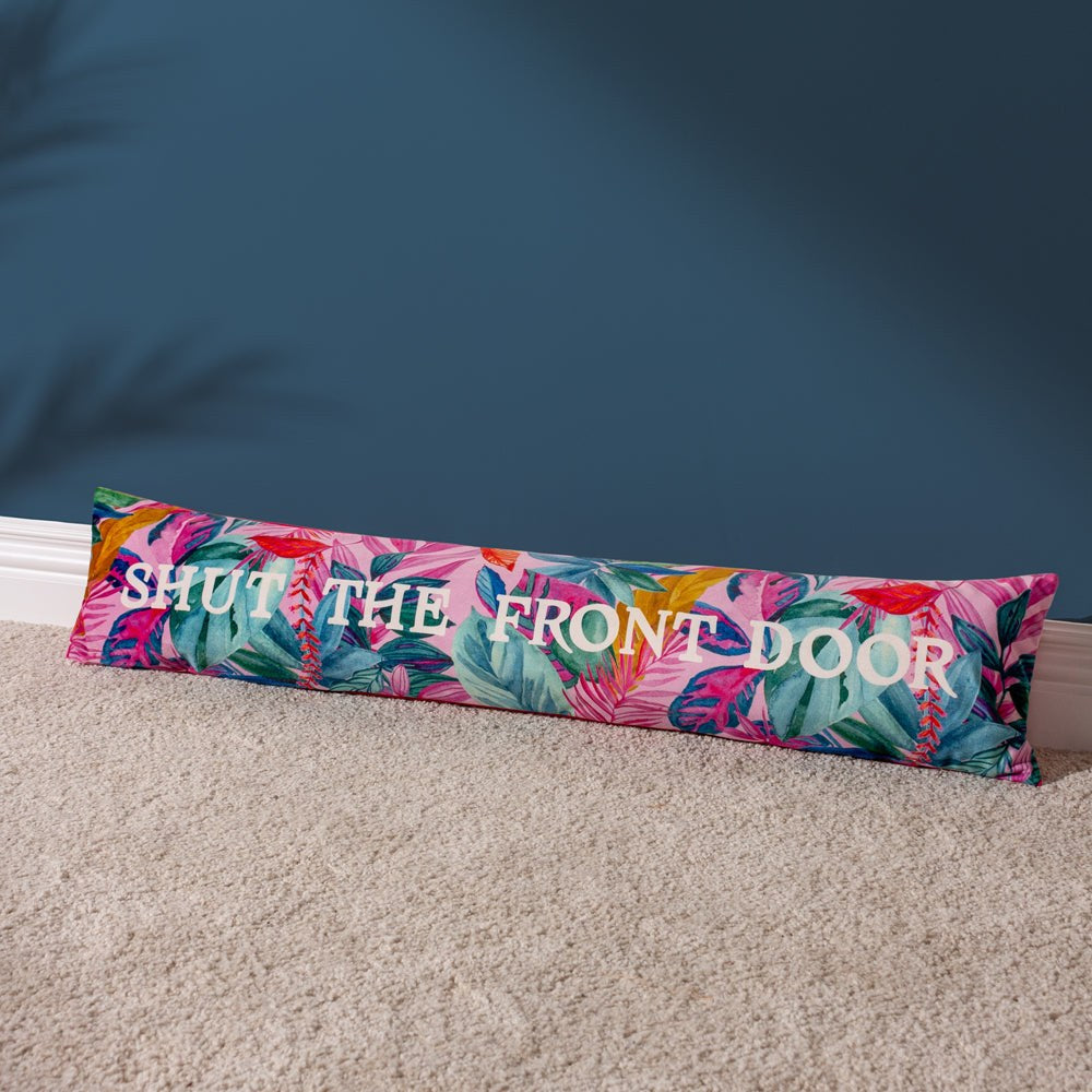 Long fuchsia velvet draught excluder printed with a tropical leaf design in pink, green and teal, bearing "Shut The Front Door" in white lettering, placed against a blue wall on carpet.