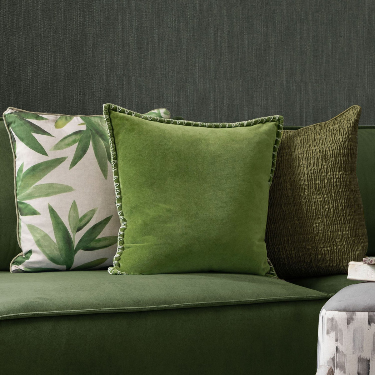 Stitch Plain Velvet Cushion in grass green with pale contrast blanket-stitch edging, shown on a green sofa beside patterned and textured cushions.