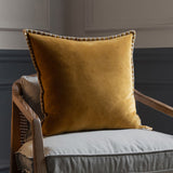 Stitch Plain Velvet Cushion in mustard yellow with contrast blanket-stitch edging, shown on a grey armchair with wooden frame.