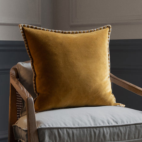 Stitch Plain Velvet Cushion in mustard yellow with contrast blanket-stitch edging, shown on a grey armchair with wooden frame.