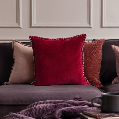 Stitch Plain Velvet Cushion in pomegranate red with pale blanket-stitch trim, shown 50x50cm on a sofa alongside blush and rust-toned cushions.