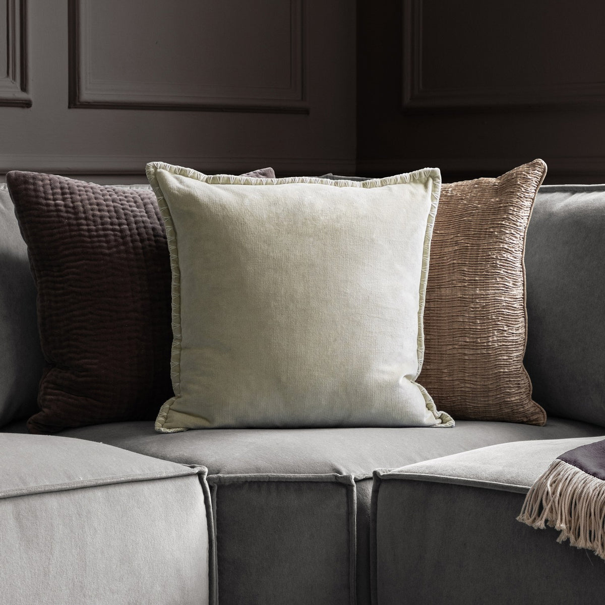Stitch Plain Velvet Cushion in Quartz grey-beige with pale cream blanket stitch trim, shown on a grey sofa with brown and taupe textured cushions.
