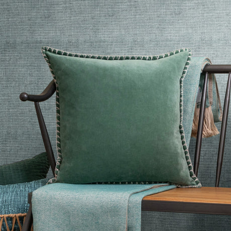 Stitch Plain Velvet Cushion in seafoam green velvet with ivory blanket-stitch edging, shown on a wooden bench with teal throws against a woven blue-grey background.