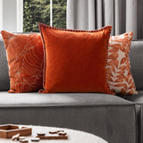 Stitch Plain Velvet Cushion in Sunset orange with contrast blanket-stitch edging, styled on a grey sofa between patterned leaf-print cushions.