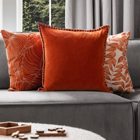Stitch Plain Velvet Cushion in Sunset orange with contrast blanket-stitch edging, styled on a grey sofa between patterned leaf-print cushions.